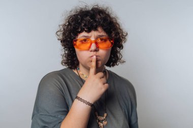 Kıvırcık saçlı, turuncu güneş gözlüğü takan ve gri gömlekli, parmakları dudaklarında şşş hareketi yapan biri. Arka plan açık gri, nötr bir atmosfer yaratıyor..