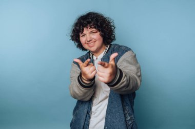 Kıvırcık saçlı, kot ceket giyen ve beyaz gömlekli, açık mavi arka planda şakacı poz veren bir genç. İki elle parmak tabancası yapıyorlar. Eğlenceli ve kendinden emin bir hava yayıyorlar..