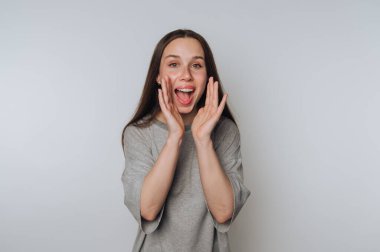 Uzun kahverengi saçlı, gri tişörtlü, neşeli bir kadın, elleriyle heyecanını ifade ediyor, yüzünü sade bir arka plana çeviriyor..