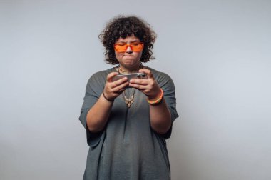 Kıvırcık saçlı, turuncu güneş gözlüğü takan, akıllı telefona odaklanmış biri..