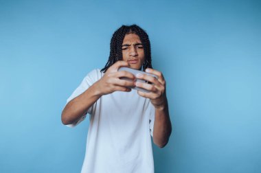 Saçları örülmüş genç bir adam mavi bir arka plana karşı konsol kumandasında yoğun bir video oyunu oynuyor..