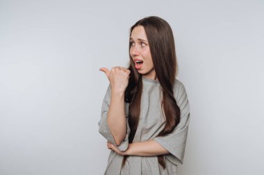 Yan tarafı gösteren uzun kahverengi saçlı, gri gömlekli, düz beyaz arka planı olan, şaşırmış genç bir kadın..