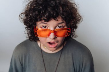 Kıvırcık saçlı, turuncu güneş gözlüklü ve günlük gömlekli biri kameraya şaşırmış bir ifadeyle bakıyor..