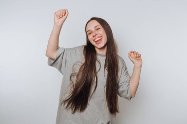 Uzun saçlı, gri tişörtlü, kollarıyla dans eden, beyaz bir arka plana karşı duran neşeli bir genç kadın..