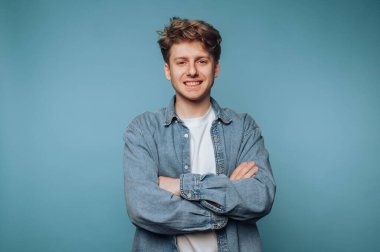 Kollarını kavuşturmuş gülümseyen genç bir adam, kot gömlek ve beyaz tişört giyiyor, mavi bir arka planı var..