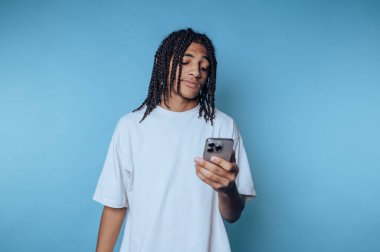 Saçları örgülü genç adam mavi arka planda akıllı telefona bakıyor..