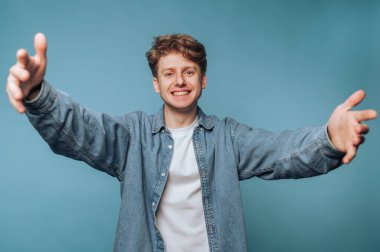 Dostça gülümseyen genç bir adam mavi bir arka plana sarılmak için kollarını uzatır..