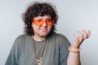 Kıvırcık saçlı, turuncu güneş gözlüğü takan ve günlük kıyafet giyen, kafa karışıklığını ifade eden biri..