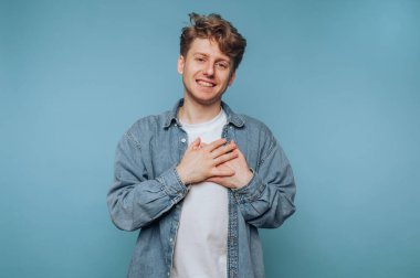 Kot gömlekli genç adam elleri göğsünde mavi arka planda gülümsüyor..