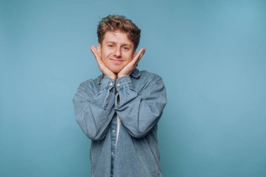 Kot gömlekli genç adam mavi arka planda şakacı poz veriyor..