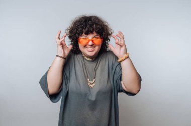 Kıvırcık saçlı, turuncu güneş gözlüğü takan, günlük giysiler giyen, gülümseyen ve ellerini arka planda gezdiren biri..