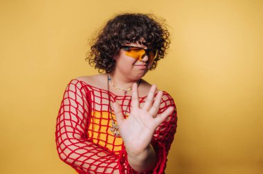 Kıvırcık saçlı, kırmızı ağ takan ve sarı arka planda güneş gözlüğü takan, eli açık hareket eden biri..