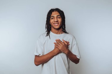 Beyaz tişört giyen, örgülü saçlı genç bir adam düz bir arkaplan karşısında duruyor, elleri göğsünün önünde birbirine kenetlenmiş gülümsüyor..