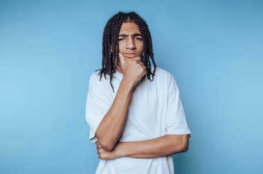 Beyaz tişörtlü genç adam, mavi arka planda düşünceli görünüyor..