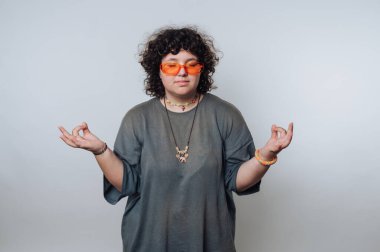 Kıvırcık saçlı, turuncu güneş gözlüklü, gri gömlekli, meditasyon yapan biri..