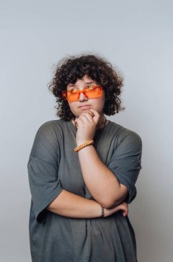 Kıvırcık saçlı, turuncu güneş gözlüklü ve gri gömlekli, düşünceli bir şekilde poz veren biri..