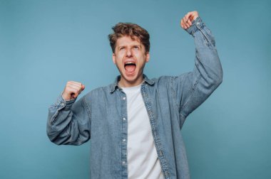 Kot gömlekli genç bir adam mavi arka planda yumruklarını kaldırarak kutluyor..