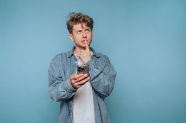Kot gömlekli genç adam mavi arka planda akıllı telefon tutarken kafa yoruyor..