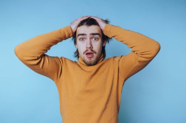 Sakallı, hardal rengi kazaklı, uzun saçlı, şaşırmış genç bir adam mavi arka planda iki eliyle başını tutuyor..