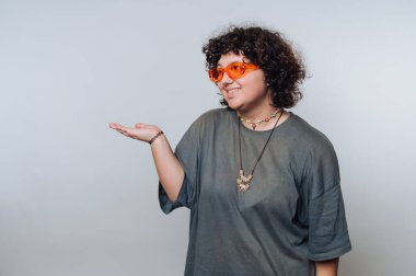 Kıvırcık saçlı, turuncu güneş gözlüğü takan ve sıradan giyinen, tek eliyle arka planda hareket eden biri..