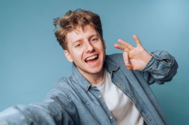 Kot gömlekli genç adam iyi bir jest yapıyor, mavi arka plana gülümsüyor..