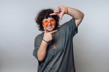 Kıvırcık saçlı, turuncu güneş gözlüğü takan ve gri tişörtlü bir insan, düz bir arkaplana karşı elleriyle yüzünü çerçeveliyor..