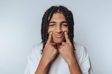 Saçları örgülü genç bir adam gülümsemek için yanaklarını çekiyor..