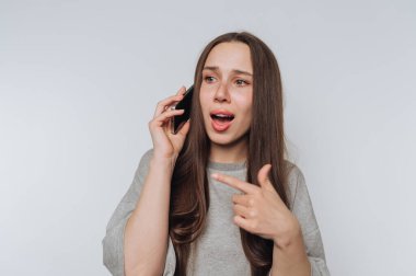 Uzun kahverengi saçlı genç bir kadın akıllı bir telefonla konuşuyor, şaşırmış ve parmağıyla işaret ediyor. Sade bir arka planda günlük gri bir bluz giyiyor..