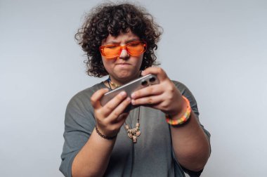 Kıvırcık saçlı, turuncu gözlüklü ve burunluk takan, akıllı telefona odaklanmış biri..