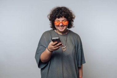 Kıvırcık saçlı, turuncu güneş gözlüklü, gri tişörtlü, düz bir arkaplanda akıllı telefona bakarken gülümseyen biri..