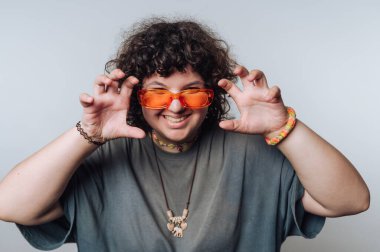 Turuncu güneş gözlüğü takan kıvırcık saçlı bir insan, pençe gibi el hareketleriyle, geniş bir gülümsemeyle şakacı poz verir. Gri bir tişört, renkli bilezikler ve kolyesi olan bir kolye takıyorlar..