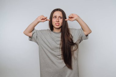 Uzun saçlı, gri bir tişört giyen, kulaklarını parmaklarıyla tıkarken sıkıntılı görünen genç bir kadın. Düz bir arkaplan üzerine kurulmuş, bunalmış ya da sıkılmış görünüyor..