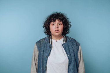 Kıvırcık saçlı, kot ceket giyen ve beyaz tişörtlü biri mavi arka planda duruyor, şaşırmış görünüyor..