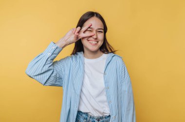 Genç bir kadın gülümsüyor ve sarı arka planda parmaklarıyla barış işareti yapıyor..