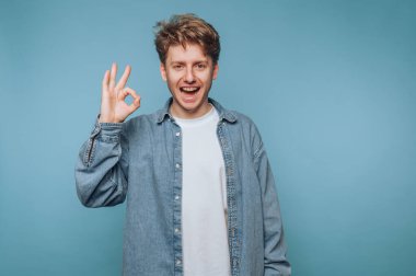 Kot gömlekli genç bir adam mavi arka plana karşı iyi bir jest yapıyor..