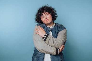 Kıvırcık saçlı bir insan mavi bir arka planda, gözleri kapalı kendini kucaklar..