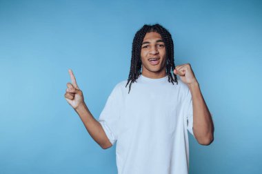 Beyaz tişörtlü, saçları örgülü genç bir adam, mavi arka planda hareketler yapıyor..