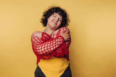 Kıvırcık saçlı biri sarılır, sarı gömleğin üzerine kırmızı bir ağ takar, sarı bir arka plana yaslanır..