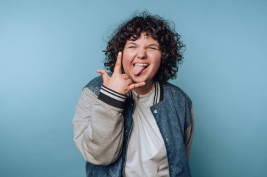 Kıvırcık saçlı, neşeli bir jest yapan, dilini dışarı çıkaran, mavi arka planda kot ceket giyen biri..