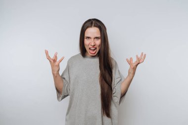 Uzun saçlı, öfkesini belli eden, bol gri gömlekli bir kadın. Yüzünde yoğun bir ifade var, ağzı açık ve elleri hayal kırıklığı içinde arka planda..