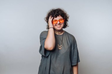 Kıvırcık saçlı, turuncu gözlüklü ve gri gömlekli gülümseyen biri..