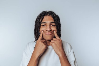 Örgülü saçlı, beyaz tişörtlü bir genç gülümserken parmaklarını kullanarak daha geniş bir tebessüm oluşturuyor..