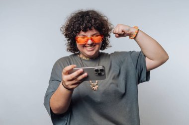 Kıvırcık saçlı, turuncu güneş gözlüğü takan ve gri tişörtlü biri akıllı telefonu tutarken gülümsüyor ve kolunu esnetiyor..