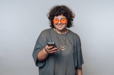 Kıvırcık saçlı, turuncu güneş gözlüklü, gri tişörtlü, düz bir arkaplanda akıllı telefona bakarken gülümseyen biri..