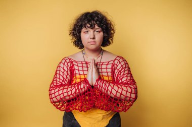 Kıvırcık saçlı, kırmızı ağlı bir insan, sarı arka plana karşı dua pozisyonunda poz verir..