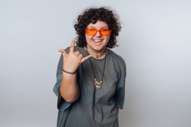 Kıvırcık saçlı, turuncu güneş gözlüklü ve gri tişörtlü biri düz bir arkaplan karşısında el hareketi yapar..