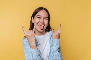 Kaya gibi el hareketleri yapan genç bir kadın, sarı bir arka plana neşeyle gülümsüyor..