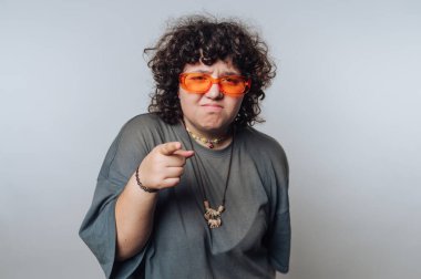 Kıvırcık saçlı, turuncu güneş gözlüklü ve gri gömlekli biri ciddi bir ifadeyle öne bakıyor..