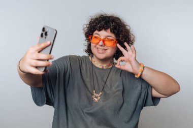 Turuncu güneş gözlüğü takan kıvırcık saçlı biri selfie çeker ve elleriyle güzel bir jest yapar. Gri bir tişört ve arka planda renkli aksesuarlar giyiyorlar..