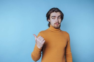 Hardallı balıkçı yakalı genç adam mavi arka planda baş parmağıyla hareket ediyor..